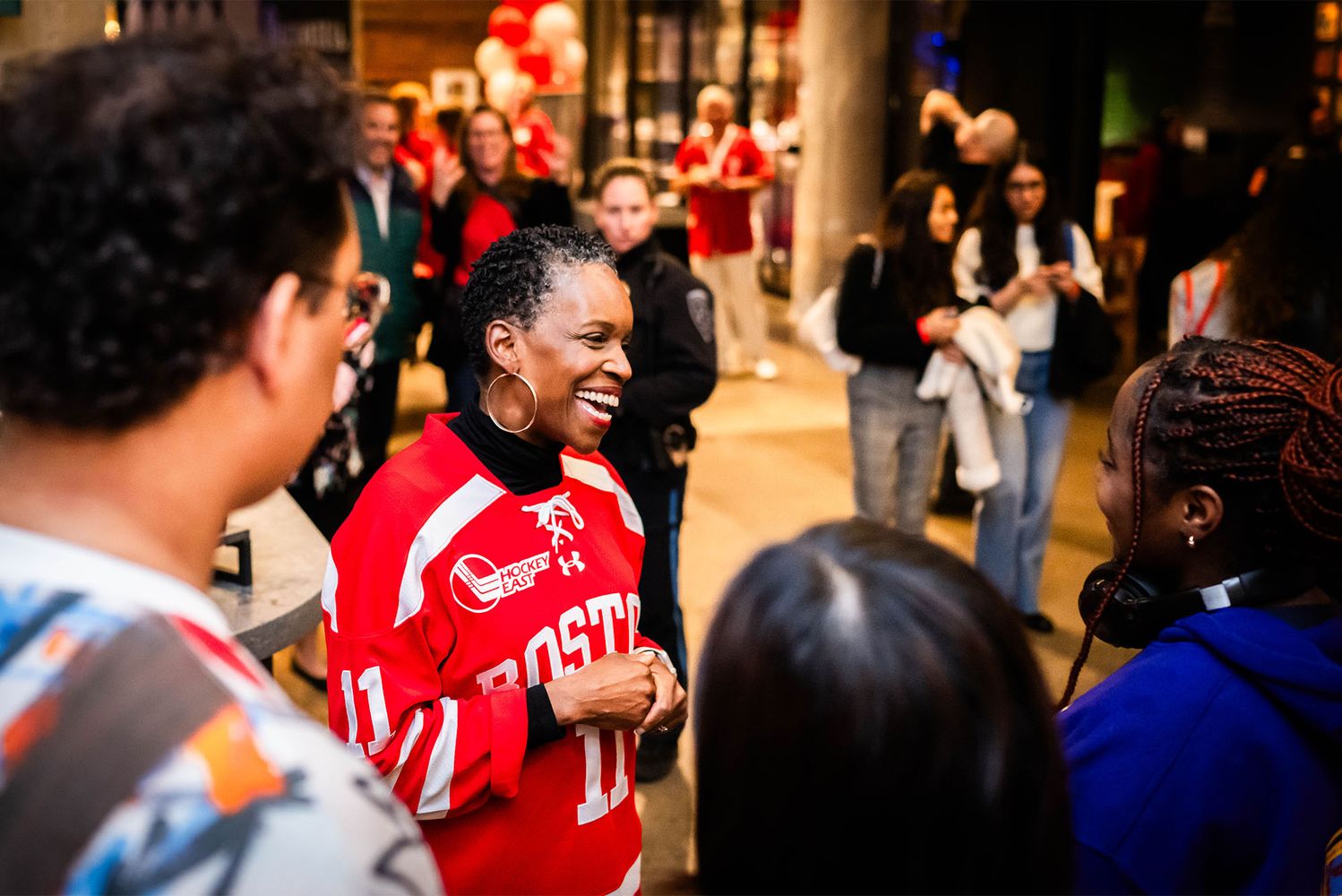 Photo: Boston University's new president Melissa Gilliam, a black woman with short dark hair in a red BU Hockey jersey, standing amongst a crowd of people with a large smile mid-conversation