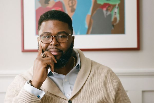 Photo: A black man with glasses sits with his chin resting on his hand in a leather chair, wearing a formal outfit in front of a piece of elegant art