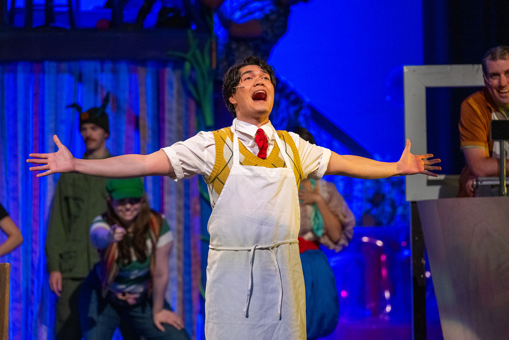 Photo: An actor onstage at the Wheelock Family Theater playing Spongebob in "Spongebob the Musical." He is in a yellow sweater vest with a white short sleeve, collared shirt and an apron. He is singing with his arms extended