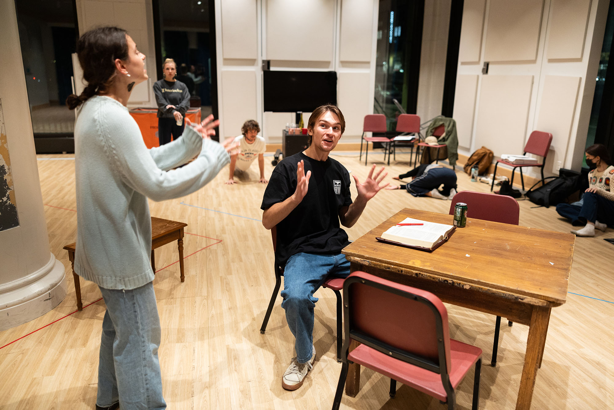 Photo: A female and male student practicing for a play. The woman, on the left, is standing with her arms raised towards the male student sitting at a table to her right.