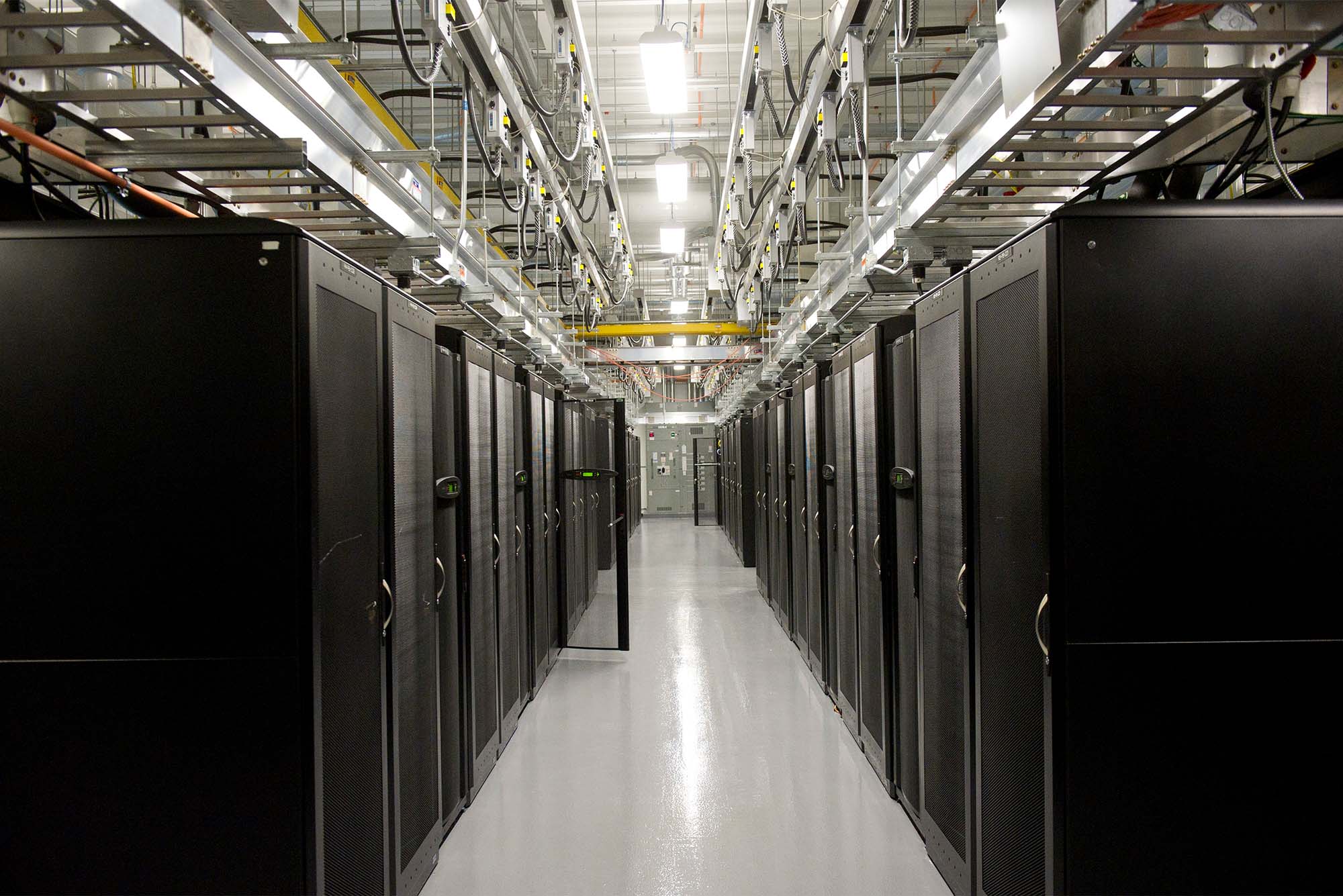 Photo: Interior of an AI data center, with large black server racks filling a gray room with steel cables and other wiring running along the ceiling