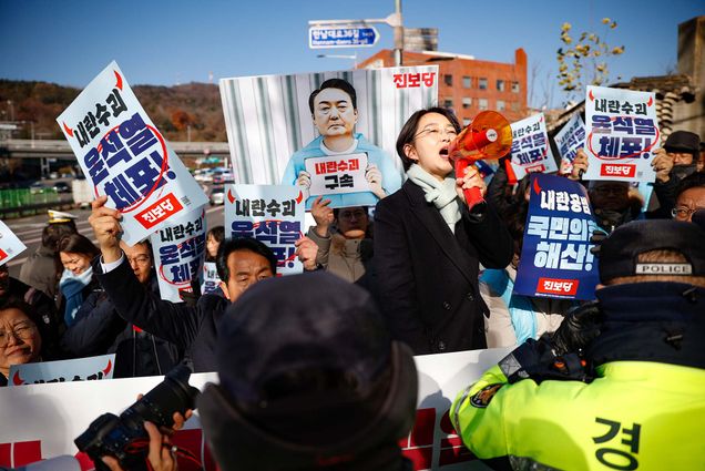 Photo: A protest in South Korea featuring a woman holding a megaphone, posters with Korean text, and a banner with an edited image of President Yoon Suk-yeol behind bars