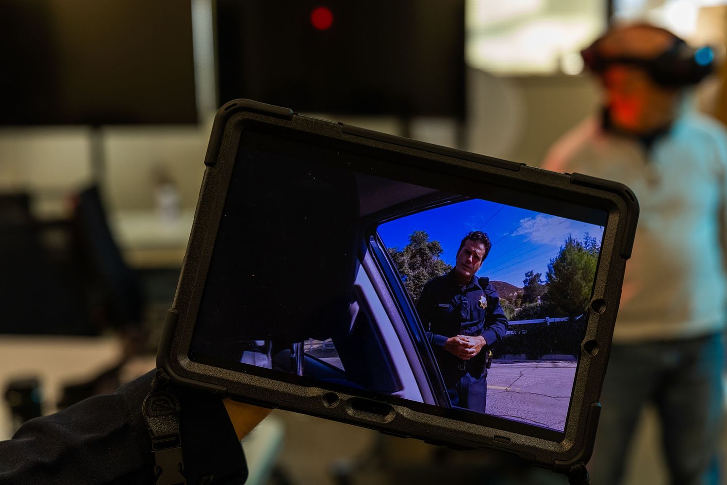 Photo: A tablet screen showing the VR simulation that a man in the background is in. It is showing an officer pulling someone over