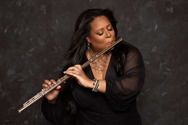 Photo: Valerie Coleman, a black woman wearing an elegant dark outfit, plays a flute in a photo studio