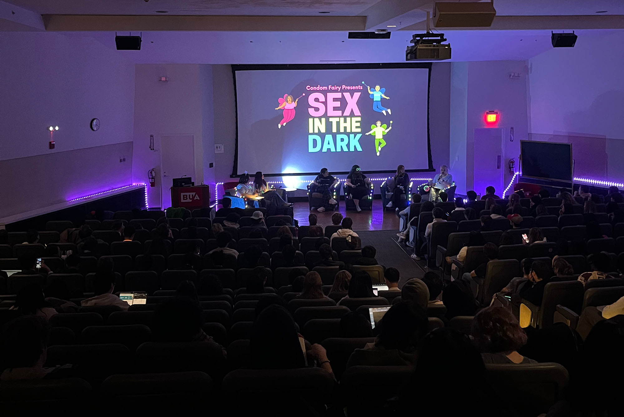 Photo: A dark room with a crowd of people facing a screen that says "Sex in the Dark"