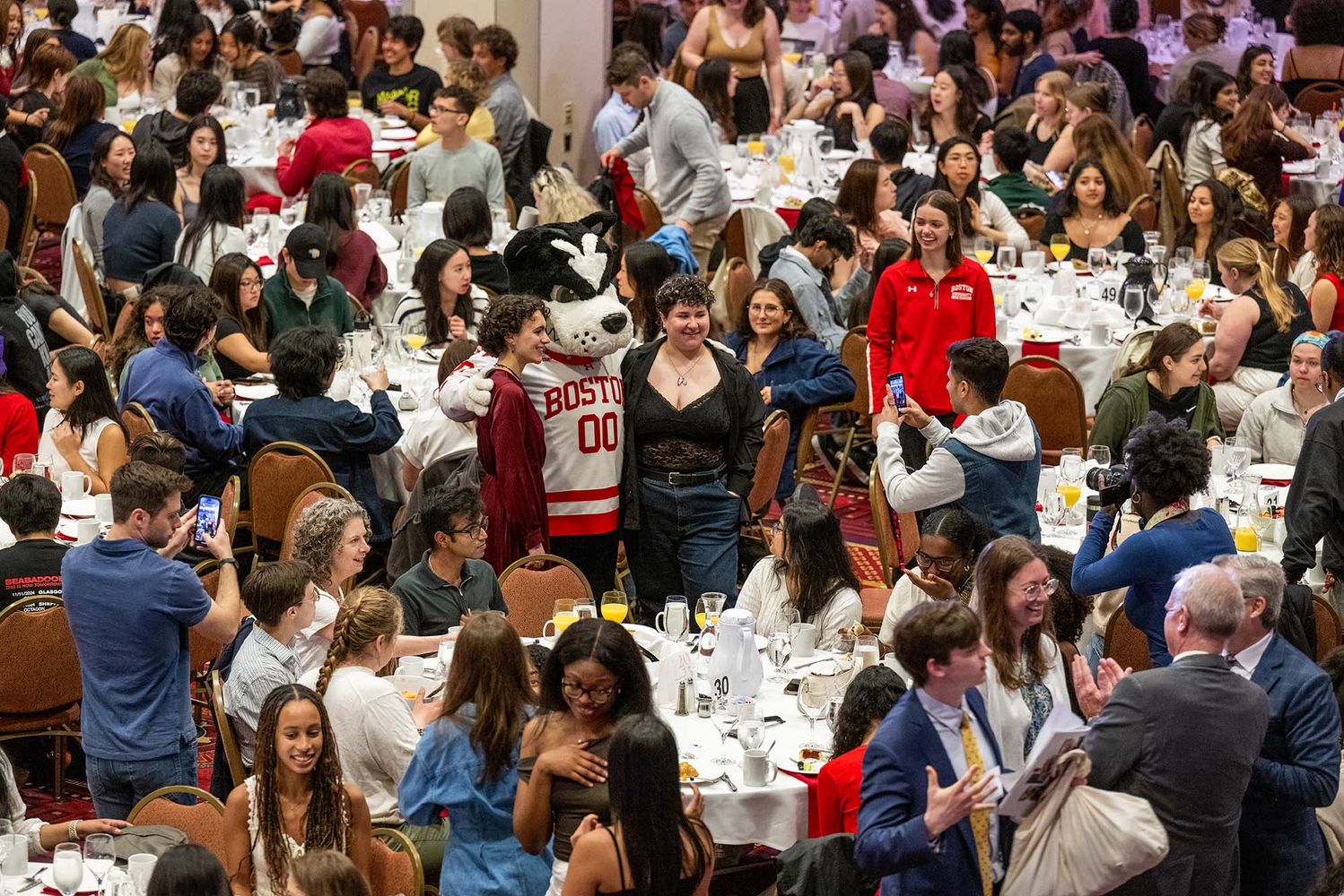 Photo: A large banquet hall filled with numerous round tables, each set with white tablecloths, plates, glasses, and cutlery. The room is crowded with people sitting at the tables and standing around. In the center of the image, a person in a Boston University mascot costume (Rhett) wearing a red and white jersey with "BOSTON 00" on it is posing for photos with attendees. Many people are taking pictures or looking at their phones. The atmosphere appears lively and social.
