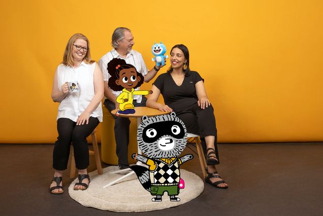 Photo: Three people smiling and posing with animations of characters from "Lyla in the Loop" and "Carl the Collector" added into the image