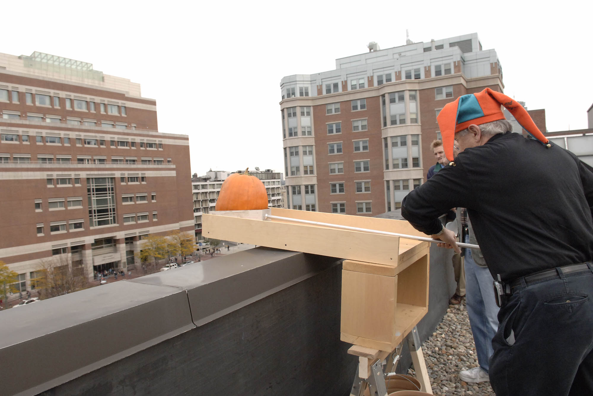 Photo: An early 2000s photo of a white man in a jester hat pushing a pumpkin off of a roof for a physics class.