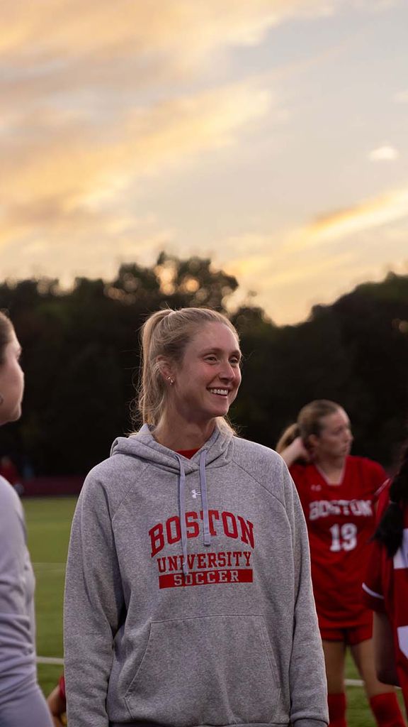 Photo: A white woman with blonde hair stands amongst players on a soccer field.