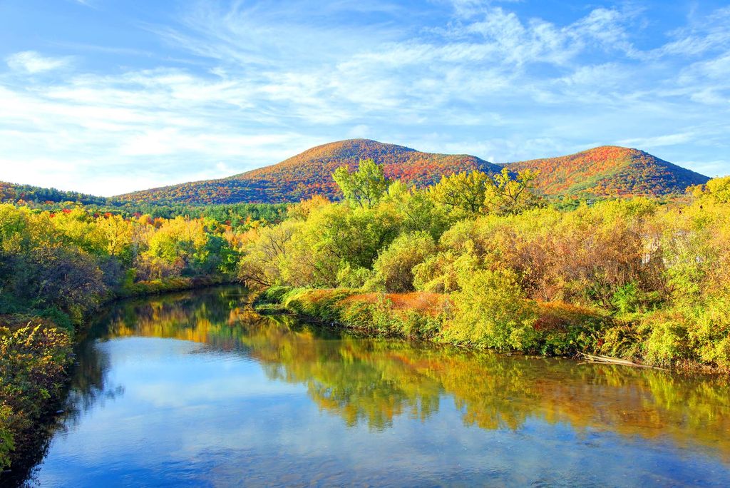 Photo: Panoramic autumn landscape with a reflective river, colorful forest, and rounded mountains beneath a blue, cloud-streaked sky.