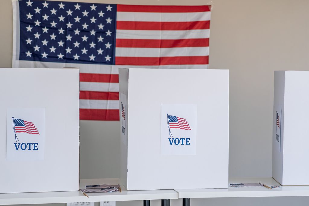 Photo: Three white voting booths labeled “VOTE” stand in front of a large American flag.