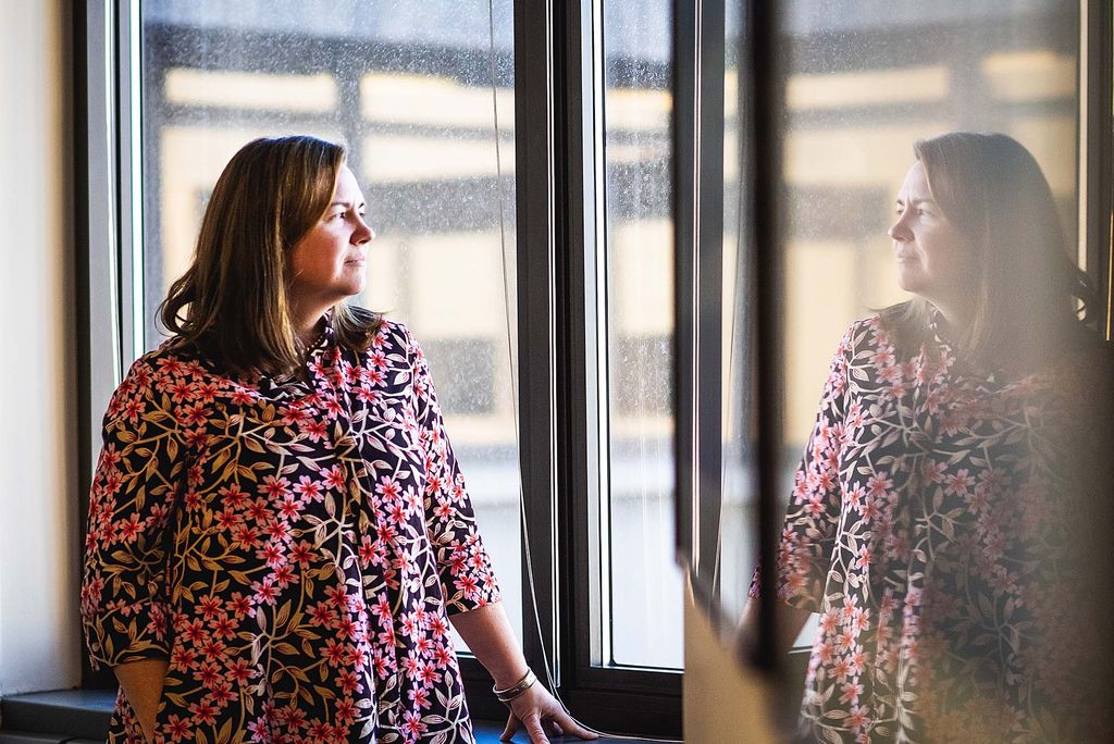 Photo: Kathryn Lamontagne gazes out the window--the photo has a double exposure making it look like she is gazing at herself. She wears a red and purple blouse with short brown hair.