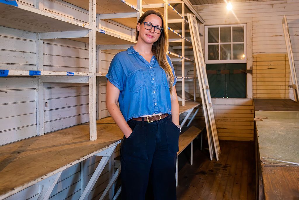 Photo: Mary Snyder, a white woman with shoulder length hair, is a BU PhD student in the American Studies program. She stands in a room with rows of wooden bare shelves.