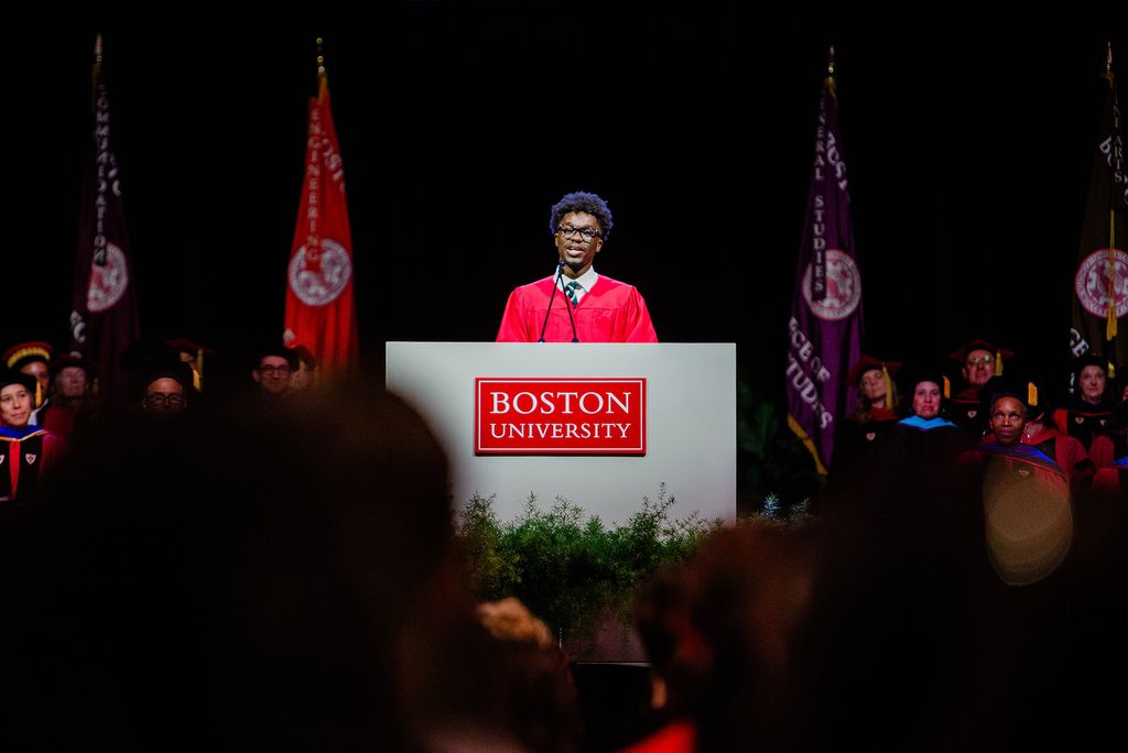 Photo: BU Student Government President Akwasi Antwi talking behind a podium that says "Boston University" while wearing a red academic gown