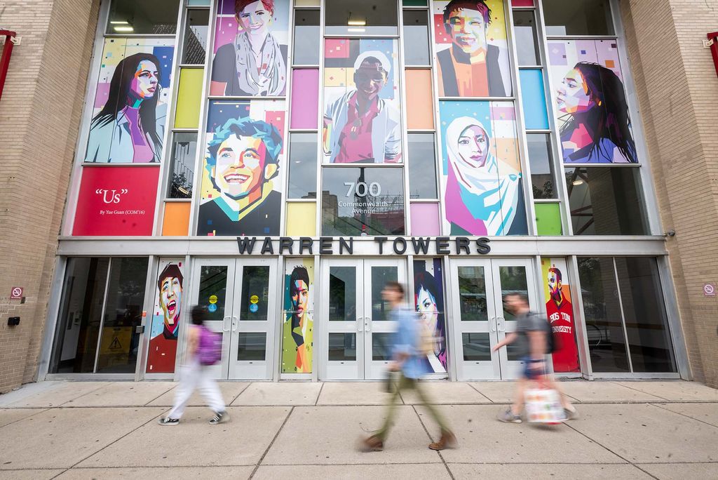 Photo: The outside of "Warren Towers," a building at BU. The outside of the building has a colorful mural of many people and there are students walking in front of the building on the sidewalk