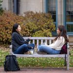 Photo: Two young women sitting cross legged on a park bench, chatting.