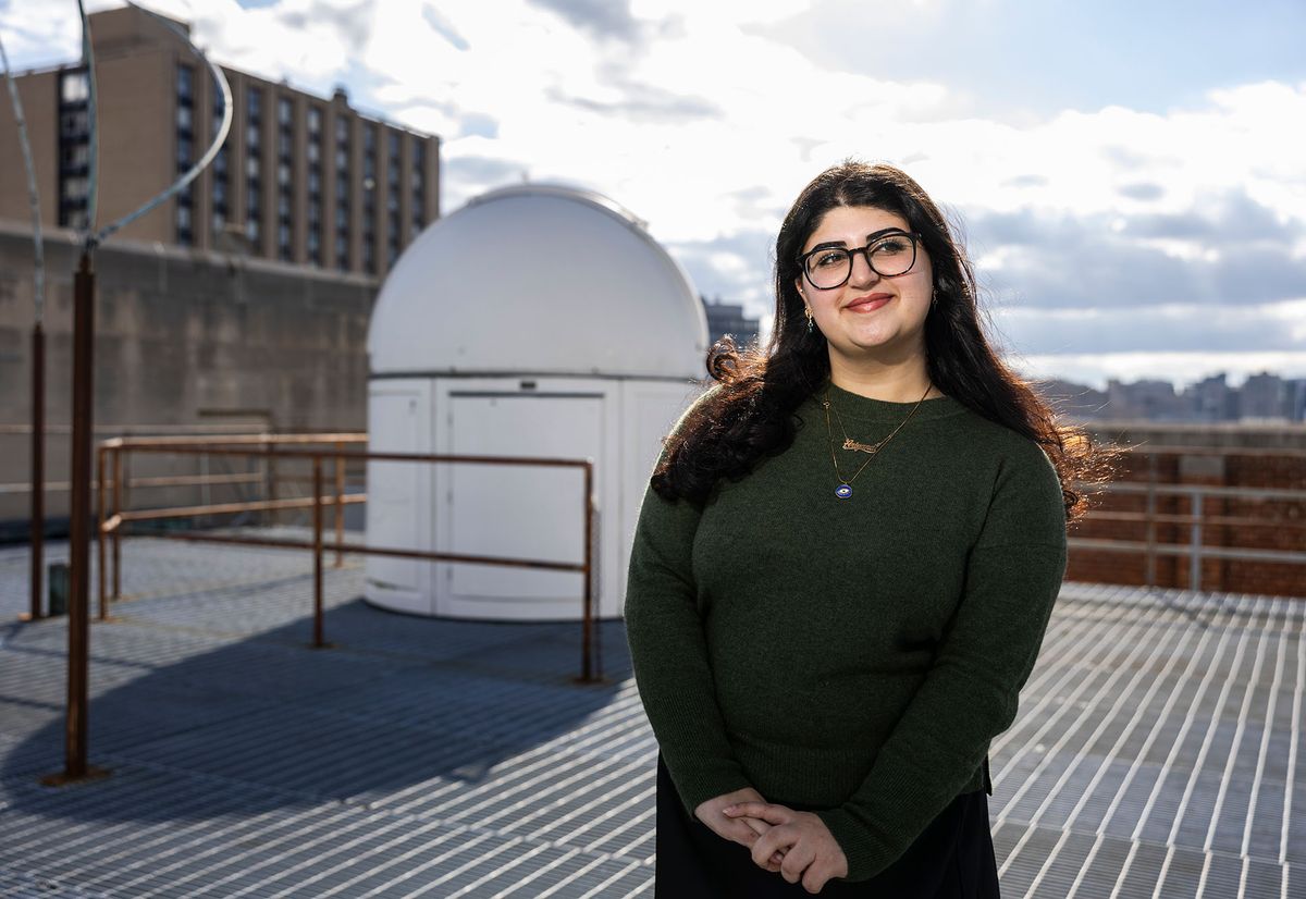 Photo: A female student standing on a rooftop with a closed smile, looking off in the distance