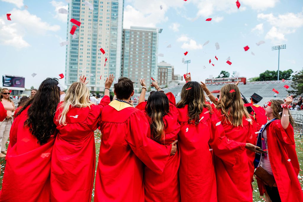 Photo: Shot of a group of graduating students wearing red gowns facing away from the camera and celebrating. Red and white confetti falls from the sky and Stuvi II can be seen in the distance.