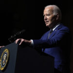 Photo: President Joe Biden speaks at an event. he stands at a podium as the right side of his face is illuminated.
