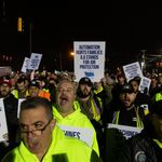 Photo: Dockworkers striking at night. They are wearing safety vests and holding signs that say "Automation Hurts Families: ILA Stands For Job Protection"