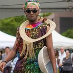 Photo: A picture of a person standing in front of a crowd while wearing colorful clothing with a fabric neck ring, hair scarf, and circular, black sunglasses. They are holding a woven basket