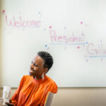 Photo: President Melissa Gilliam sits underneath a whiteboard with a handwritten note reading "Welcome President Gilliam"