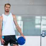 Photo: A muscular young man holding a blue basketball stands next to an iPhone on a tripod in a gym environment