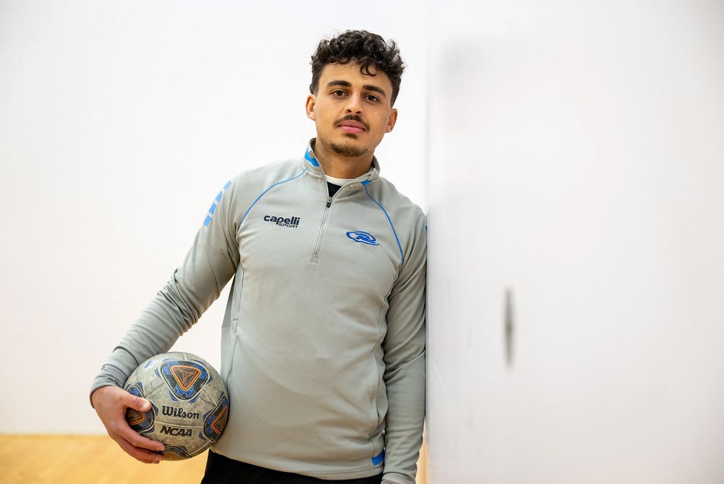 Photo: A young man with short dark hair holding a soccer ball leaning against a wall