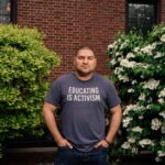 Photo: A man in a gray t shirt with text reading "Education is activism" stands in front of a brick wall with a stoic expression