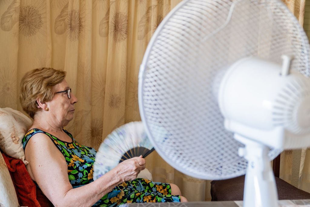 Photo: A picture of an older woman sitting on the sofa and cooling herself with a paper fan and an electric fan