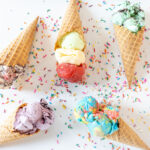 Photo: A white table top with five different ice creams with a waffle cone are scatterd in a flat-lay.