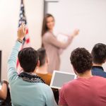 Photo: A student raising his hand during a class. His female teacher is turning to look back at him and stands at the front of a whiteboard next to an American flag