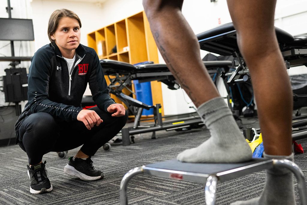 Photo: A female athletic trainer kneeling and watching as an athlete does a stepping exercise