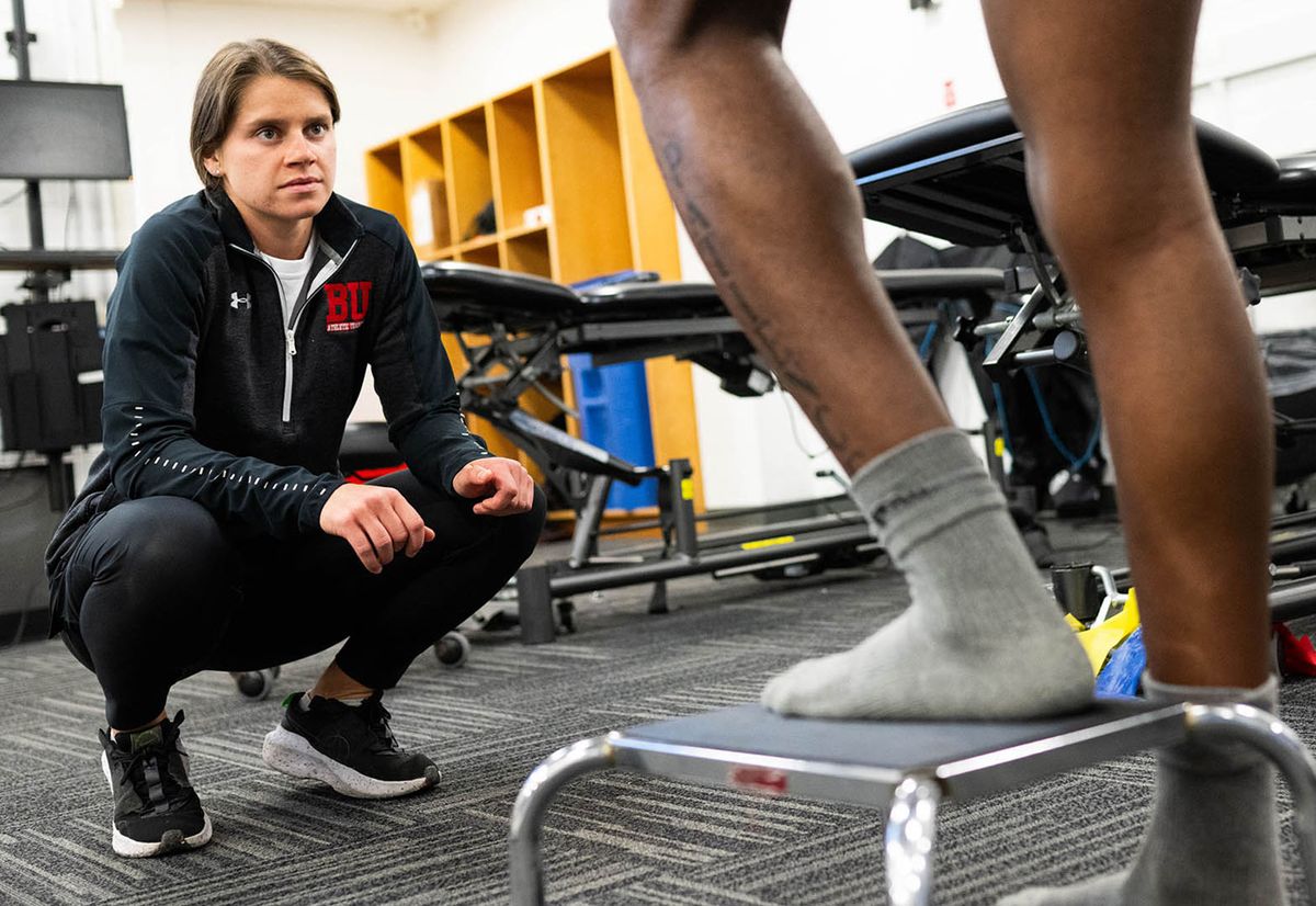 Photo: A female athletic trainer kneeling and watching as an athlete does a stepping exercise