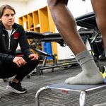 Photo: A female athletic trainer kneeling and watching as an athlete does a stepping exercise