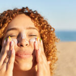 Photo: A picture of a smiling woman with curly hair applying sunscreen to her face on the beach
