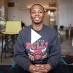 Photo: A young Black man sitting in a chair with a cross around his neck and a Chicago Bulls sweatshirt. He smiles for the photo. Video play overlay