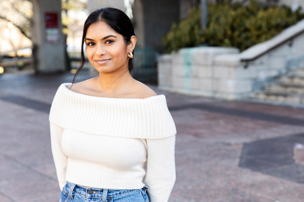 Photo: Sindhu Rayapaneni, a young woman with long dark hair wearing an off-the-shoulder cream sweater, poses for a photo on Marsh Plaza.