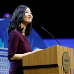 Photo: A picture of Boston Mayor Michelle Wu standing behind a podium. She is wearing a quarter-sleeve burgundy dress and smiling while looking into the crowd