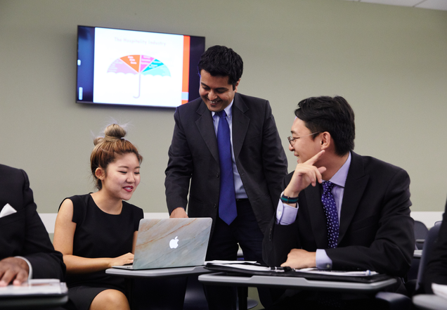 Two men and a woman interacting in a classroom setting. The woman and one man are sitting, while the other man is standing. They are professionally dressed and all directing their attention to the woman's laptop screen.