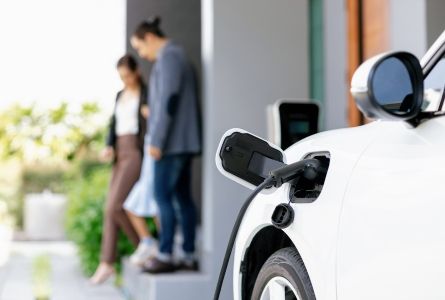 Focus closeup electric vehicle recharging battery from home electric charging station with blurred family in background.