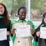 Three BU students posing in green capes holding award certificates.