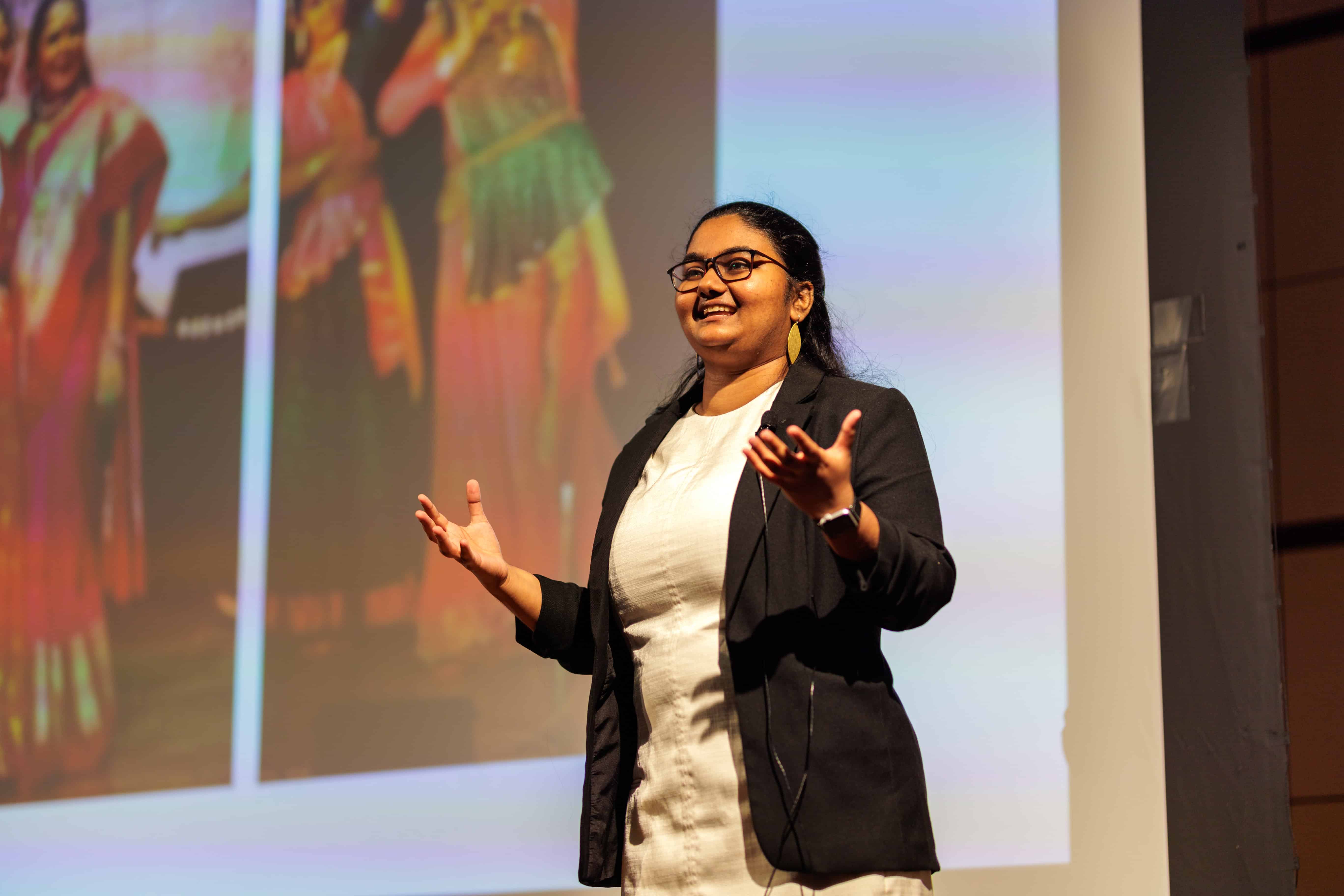 Woman presents on a stage in front of a projector screen while gesturing with her hands