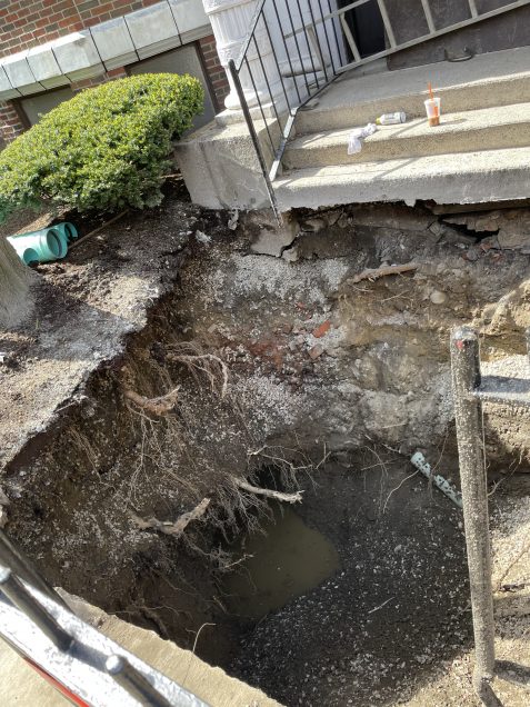 A large work site hole in front of a building with tree routes protruding into the hole.