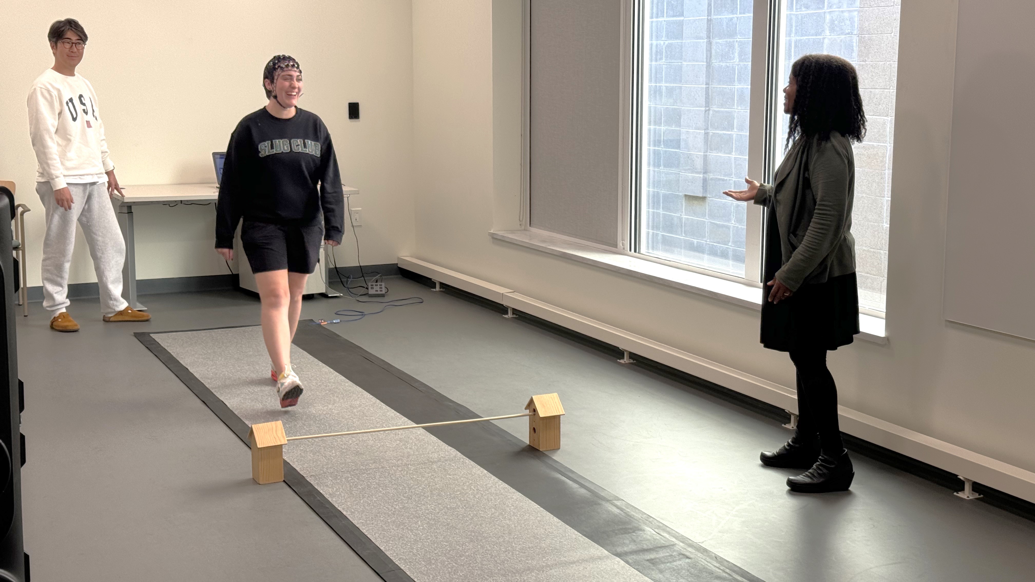 Three people in a lab environment, one of them is walking across a mat and wearing experimental headgear