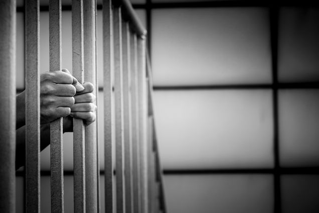 A black and white image of hands clutching the bars of a prison cell