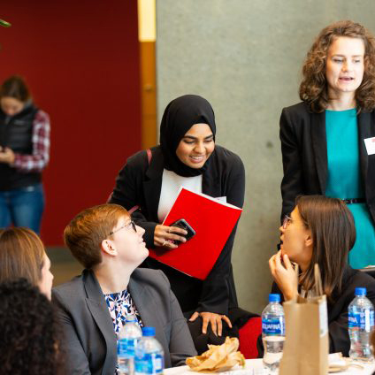 Student in hijab holding a red folder leans over desk at LAW MarComm 1L Career Conference to talk to fellow students