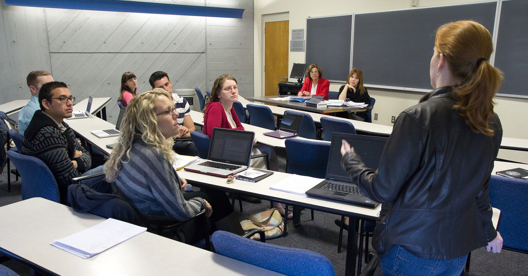 Several students sit at rows of tables and discuss a case in the foreground. Two professors sit at a table in the background.