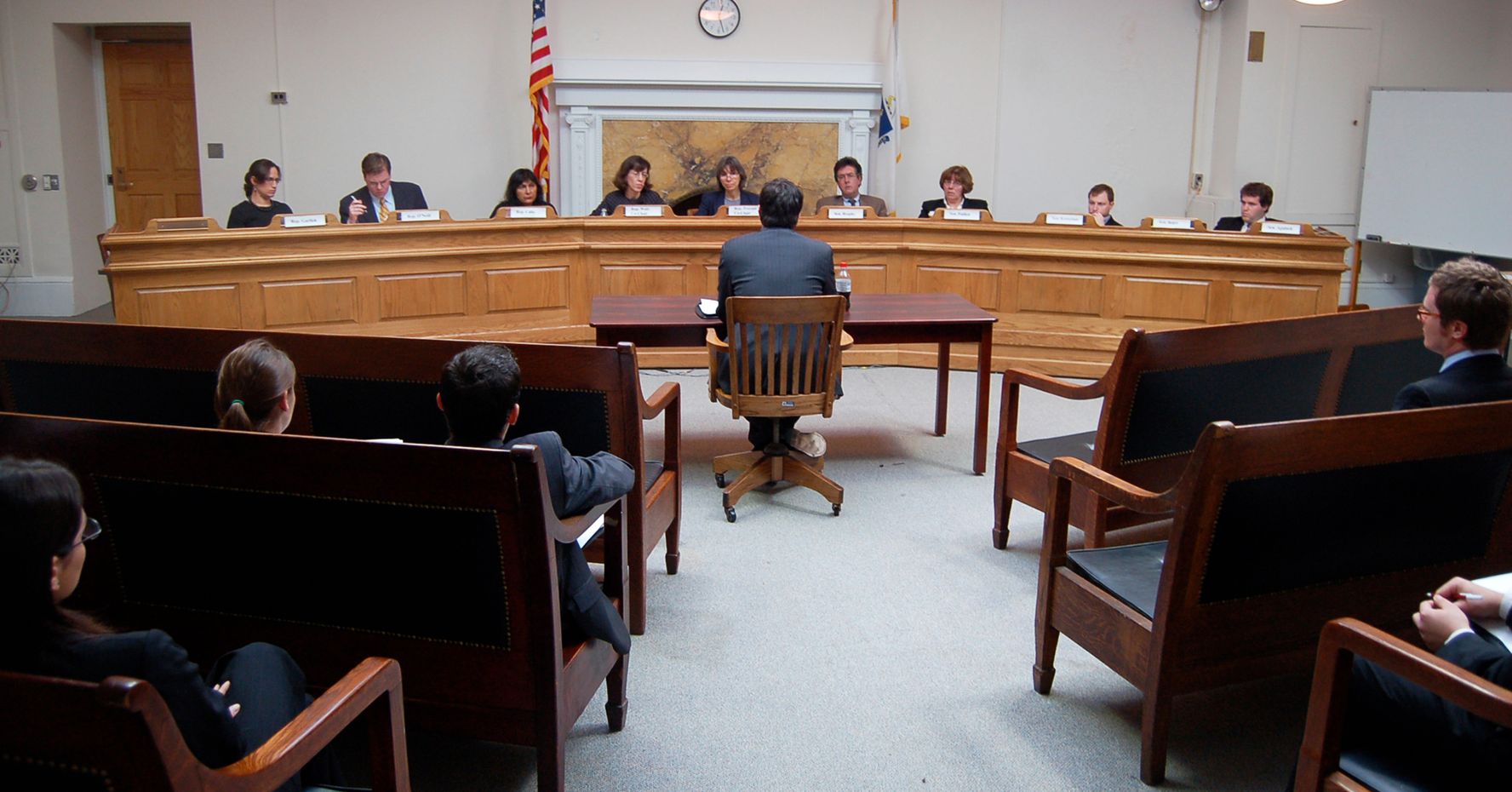 A student sits in a large courtroom in front of a panel of judges. Other students observe in the foreground.