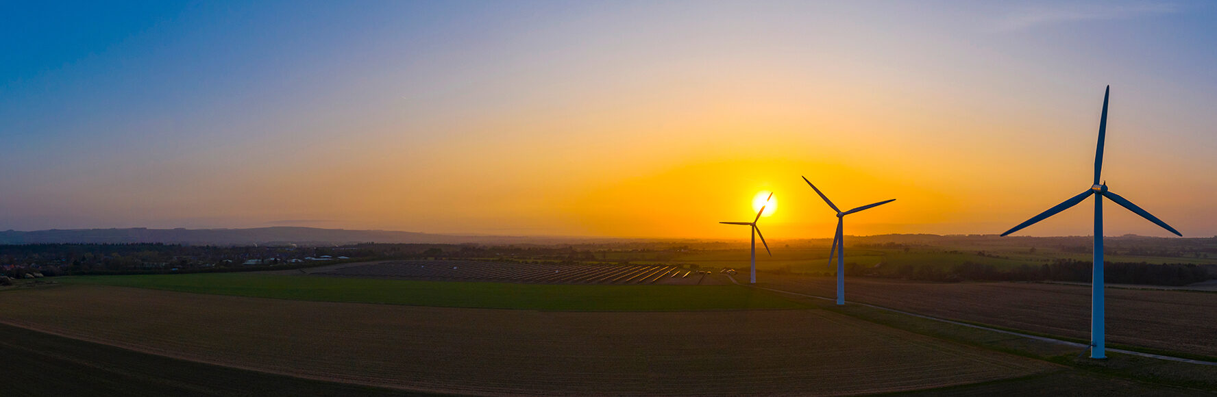 Modern windmills loom over a field of crops at sunset.